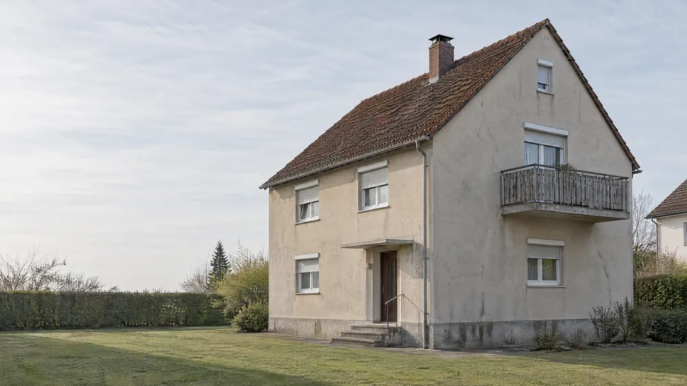 Konzeptvisualisierung, Zustand Bestand: zweigeschossiges Wohnhaus mit gealtertem beigegrauem Putz, Rissen, weißen Kunststofffenstern mit Rollläden und einem Holzbalkon.